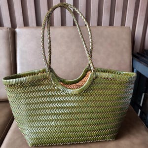 Puede incluir: Un bolso tote verde tejido con asas trenzadas. El bolso tiene forma rectangular con una abertura ancha y un patrón tejido. Las asas son de un tono verde más oscuro. El bolso está sobre una superficie marrón.