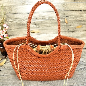 K&ouml;nnte beinhalten: Eine braune, gewebte Tragetasche mit geflochtenem Griff. Die Tasche hat eine breite &Ouml;ffnung und einen Kordelzugverschluss. Die Tasche besteht aus einem gewebten Material und hat ein nat&uuml;rliches, rustikales Aussehen. Die Tasche ist zum Tragen von Gegenst&auml;nden bestimmt.