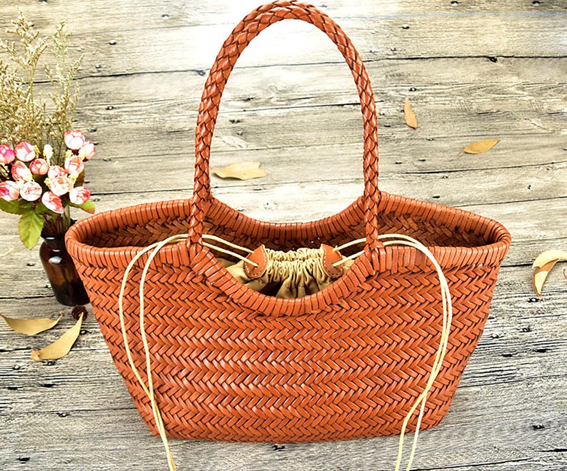 K&ouml;nnte beinhalten: Eine braune, gewebte Tragetasche mit geflochtenem Griff. Die Tasche hat eine breite &Ouml;ffnung und einen Kordelzugverschluss. Die Tasche besteht aus einem gewebten Material und hat ein nat&uuml;rliches, rustikales Aussehen. Die Tasche ist zum Tragen von Gegenst&auml;nden bestimmt.