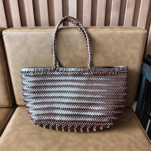 Può includere: Una borsa tote marrone intrecciata con manici intrecciati. La borsa presenta un motivo a spina di pesce orizzontale ed è realizzata in un materiale marrone scuro. La borsa è appoggiata su una superficie in pelle marrone.