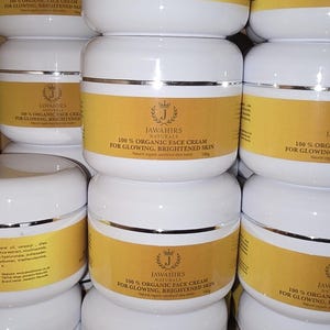 Könnte beinhalten: Gestapelte weiße Tiegel Gesichtscreme mit goldenen Etiketten. Auf den Etiketten steht "100% Organic Face Cream for Glowing, Brightened Skin". Der Markenname "Jawahirs Naturals" ist ebenfalls zu sehen. Die Tiegel sind übereinander gestapelt.