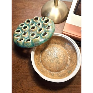 May include: A ceramic trinket dish with a lid. The lid has a green and brown glaze and features a design of multiple circular holes. The dish is glazed in a brown and white speckled pattern.