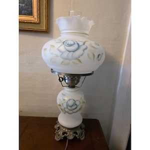 May include: A white, vintage-style table lamp with a floral design. The lamp features a frosted glass shade and base, adorned with blue rose motifs and green leaves. The base is ornate, with a silver-colored metal finish.