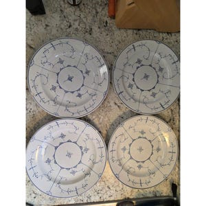 May include: Four white ceramic plates with a blue floral pattern. Each plate has a central floral design encircled by fine blue lines and dots. The plates are arranged on a light-coloured countertop.