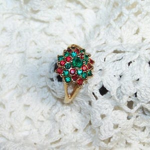 May include: A gold-toned ring with a cluster of red and green gemstones surrounding a central green stone. The ring has a floral design and is set against a white, crocheted background.
