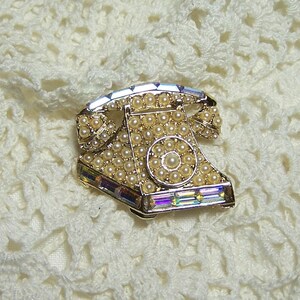May include: A gold-tone brooch shaped like a vintage rotary phone, decorated with iridescent rhinestones and faux pearls.