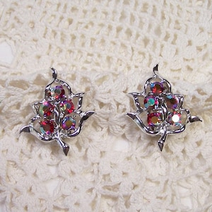 May include: A pair of silver-toned leaf-shaped earrings. Each earring features a cluster of iridescent, red, and purple gemstones. The earrings are set against a cream-colored, crocheted background.