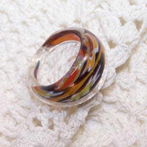 May include: A clear glass ring with colorful swirls of orange, brown, yellow, and black. The ring is set against a white, crocheted background, showcasing the intricate floral pattern of the fabric.