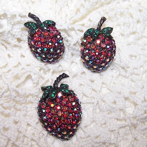 May include: Three red and green rhinestone strawberry brooches. The brooches are made of metal and have a shiny finish.