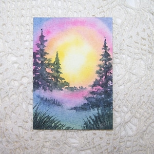 May include: Watercolor painting of a sunset over a forest. The sky transitions from pink to yellow, with silhouetted evergreen trees. Green grass and blue water are in the foreground. The artwork is on a rectangular piece of paper.