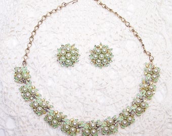 May include: A vintage jewelry set featuring a necklace and matching earrings. The necklace and earrings have a floral design with faux pearls, pale green enamel, and small clear rhinestones. The set is set on a gold-tone metal.