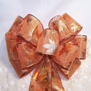 May include: A large, decorative bow made of orange ribbon with a leaf pattern and metallic gold accents. The bow has a wired edge and is suitable for fall or autumn-themed decorations.