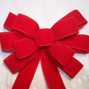 May include: A vibrant red velvet bow with a decorative design. The bow is centered and has multiple loops and tails. The edges are finished with a matching red trim. The bow is set against a white background.