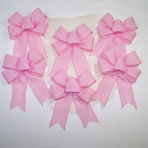 May include: Six light pink fabric bows with long tails are arranged on a white crocheted doily. The bows are evenly spaced and have a classic bow shape. The background is a plain white surface.