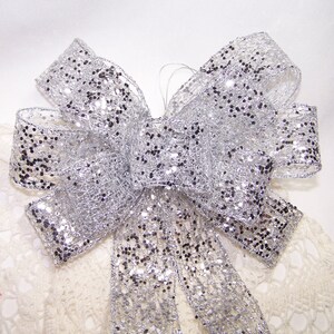 May include: A silver glitter bow with a mesh design. The bow is tied and has multiple loops and streamers. The glitter is black and silver, and the bow is set against a white background.