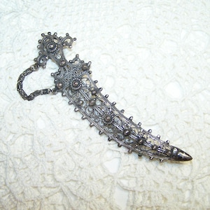 May include: Ornate silver-toned dagger with intricate filigree detailing. The handle and blade feature floral and circular embellishments. A delicate chain is attached to the handle. The dagger rests on a white, textured surface.