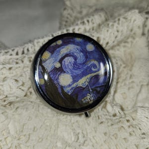 May include: A round, silver-toned metal pill box featuring a glossy, domed top printed with a reproduction of Vincent van Gogh's The Starry Night painting. The artwork displays swirling blue and yellow patterns, a dark cypress tree, and a village.