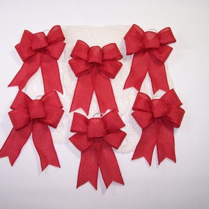 May include: Six red fabric bows with long, pointed tails are arranged on a white lace background. The bows are made of a sheer material, and their color is a deep, vibrant red. The bows are evenly spaced.