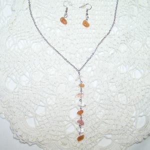 May include: A silver-toned necklace and matching earrings set. The necklace features a vertical strand of orange, pink, and clear gemstones. The earrings each have a single orange gemstone. The jewelry is displayed on a white, lacy surface.