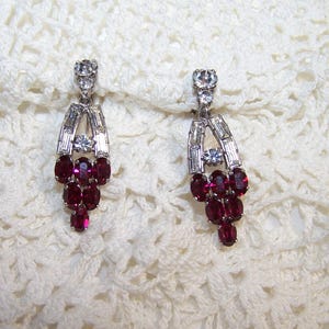 May include: A pair of drop earrings featuring a cluster of oval-cut deep red stones at the base, accented by clear baguette and round-cut stones set in a silver-toned metal frame, with a single round clear stone at the top of each earring.
