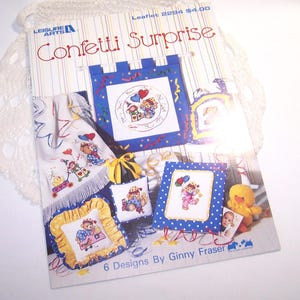 May include: A craft leaflet titled "Confetti Surprise" featuring cross-stitch designs. The cover displays several finished projects with teddy bears and other festive elements. The designs are framed in blue and yellow, with a white lace trim.