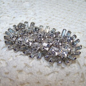 May include: A silver-toned brooch with a cluster of clear, sparkling rhinestones. The brooch has an oval shape with a central arrangement of square-cut stones and elongated, rectangular stones radiating outwards.