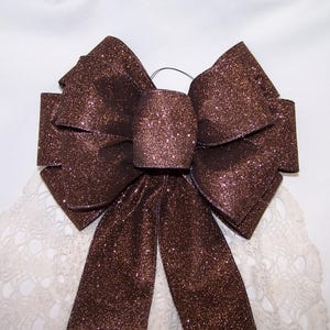 May include: A decorative bow crafted from dark brown fabric with a dense, shimmering glitter finish. The bow features multiple structured loops and two long, hanging tails extending downward, creating a full and symmetrical appearance.