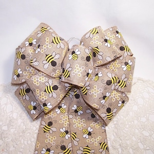 May include: A large decorative bow made of tan burlap ribbon. The ribbon is printed with a pattern of yellow and black bees and honeycomb shapes. The bow is tied and has multiple loops and tails, suitable for wreaths or other crafts.
