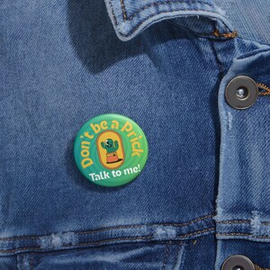 May include: A round, green button with the text "Don't be a Prick" and "Talk to me!" in a playful font. The button features a cartoon cactus in a pot. The button is pinned to a blue denim jacket.