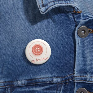 May include: A white button with a red button in the center. The button has four holes. The text "up for love" is printed below the button.