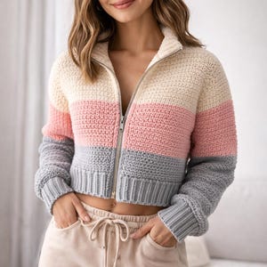 May include: A cropped, color-blocked crochet jacket with a zip-up front. The jacket features horizontal stripes of cream, pink, and gray. The model is wearing beige sweatpants. The jacket has long sleeves and a ribbed hem.