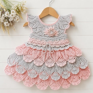 May include: A handmade crochet dress in gray and pink, hanging on a wooden hanger. The dress features a scalloped design with a flower embellishment at the neckline. The dress is approximately 24 inches long.