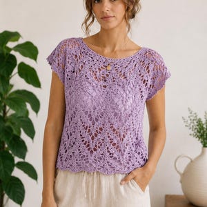 Puede incluir: Top de ganchillo lila con mangas cortas y diseño calado. El top, con cuello redondo, se combina con pantalones beige. El fondo muestra una planta y un jarrón. Es una prenda delicada, hecha a mano.