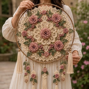 May include: A circular wall hanging featuring a cream-colored crocheted lace doily base adorned with multiple three-dimensional dusty rose flowers and dark green crocheted leaves. The hoop includes hanging cream tassels and floral accents.