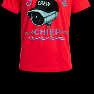 May include: A bright red short-sleeved t-shirt featuring a graphic of a security camera wearing a black baseball cap with the word CREW. Above the camera is the text CAMERA CREW, and below is the word CHIEF with two keys and wavy lines.
