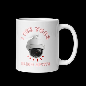 May include: A white ceramic mug featuring a graphic of a white dome security camera in the center. Above the camera, the red text reads I SEE YOUR, and below the camera, the red text reads BLIND SPOTS, with small red lines radiating from the lens.