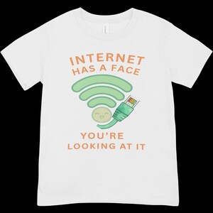 以下が含まれることがあります： 笑顔のWi-Fiシンボルとイーサネットケーブルコネクタのグラフィックが描かれた白い半袖Tシャツ。グラフィックの上下には、オレンジ色の文字でINTERNET HAS A FACE YOU'RE LOOKING AT ITと書かれています。