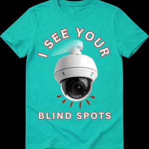 May include: A bright teal short-sleeve t-shirt featuring a white dome security camera graphic in the center. The text "I SEE YOUR" is arched above the camera, and "BLIND SPOTS" is printed below in white capital letters with a red outline.