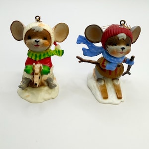 May include: Two mouse ornaments with hanging loops. One mouse wears a white hat, red coat, and green scarf while riding a rocking horse. The other mouse wears a red knit hat and blue polka-dot scarf while skiing on wooden skis with poles.