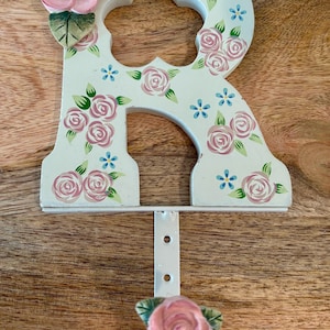 Peut inclure: Lettre "R" décorative en bois, ornée de motifs floraux. Peinte en blanc, elle présente des roses roses, des feuilles vertes et des fleurs bleues. Un crochet en métal blanc avec une rose est fixé en bas. Décoration murale.