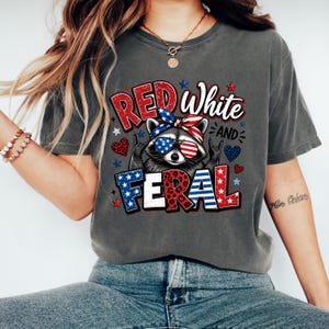 Puede incluir: Camiseta gris oscuro con un mapache con gafas de sol y una bandana roja, blanca y azul. La camiseta muestra las palabras "Red White and Feral" en una fuente patri&oacute;tica, con estrellas y corazones.