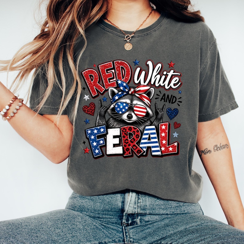Puede incluir: Camiseta gris oscuro con un mapache con gafas de sol y una bandana roja, blanca y azul. La camiseta muestra las palabras "Red White and Feral" en una fuente patri&oacute;tica, con estrellas y corazones.