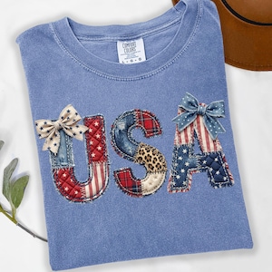 May include: A light blue t-shirt with the word "USA" appliqued on the front. The letters are made of patchwork fabric in red, white, blue, and leopard print patterns, with decorative bows. The shirt has a crew neck and a tag that says "Comfort Colors".
