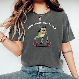 Puede incluir: Camiseta gris oscuro con un dibujo de una rana de dibujos animados con un sombrero de bruja en un patinete. El texto "off to commit aunt foolery" está encima de la rana. La rana es verde y beige, el patinete es negro y rosa.