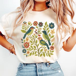 May include: Cream-colored t-shirt featuring a colorful illustration of three birds perched among flowers and foliage. The birds have blue, green, and orange coloring. The flowers are in shades of red, yellow, and blue.