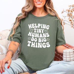 May include: An olive green t-shirt featuring the white text "HELPING TINY HUMANS DO BIG THINGS", with heart accents. The t-shirt has a relaxed fit and is a casual style.