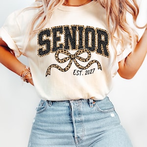 May include: A cream-colored t-shirt featuring the word SENIOR in black glittery letters with a leopard print outline. Below the text is a large leopard print bow graphic and the black text EST. 2027. The shirt is paired with light blue denim jeans.