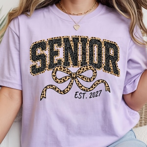 May include: A light purple t-shirt featuring the word SENIOR in black glittery letters with a leopard print outline. Below the text is a large leopard print bow graphic and the black text EST. 2027. The shirt has a crew neckline and short sleeves.