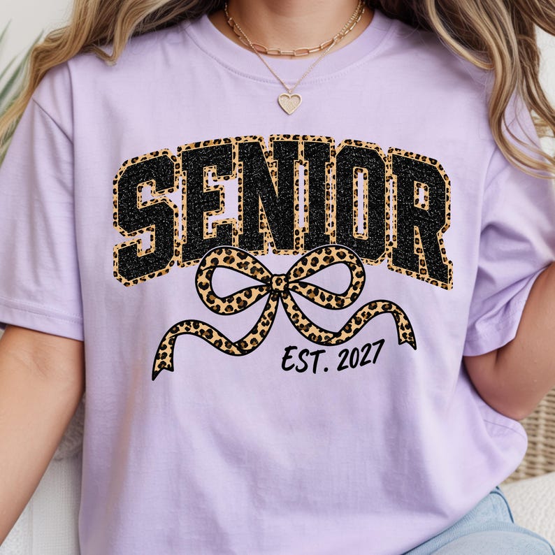 May include: A light purple t-shirt featuring the word SENIOR in black glittery letters with a leopard print outline. Below the text is a large leopard print bow graphic and the black text EST. 2027. The shirt has a crew neckline and short sleeves.