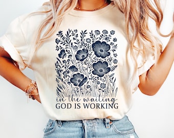 May include: Cream-colored t-shirt featuring a navy blue floral design with the text "in the waiting GOD IS WORKING". The graphic includes stylized flowers and foliage, creating a botanical theme.